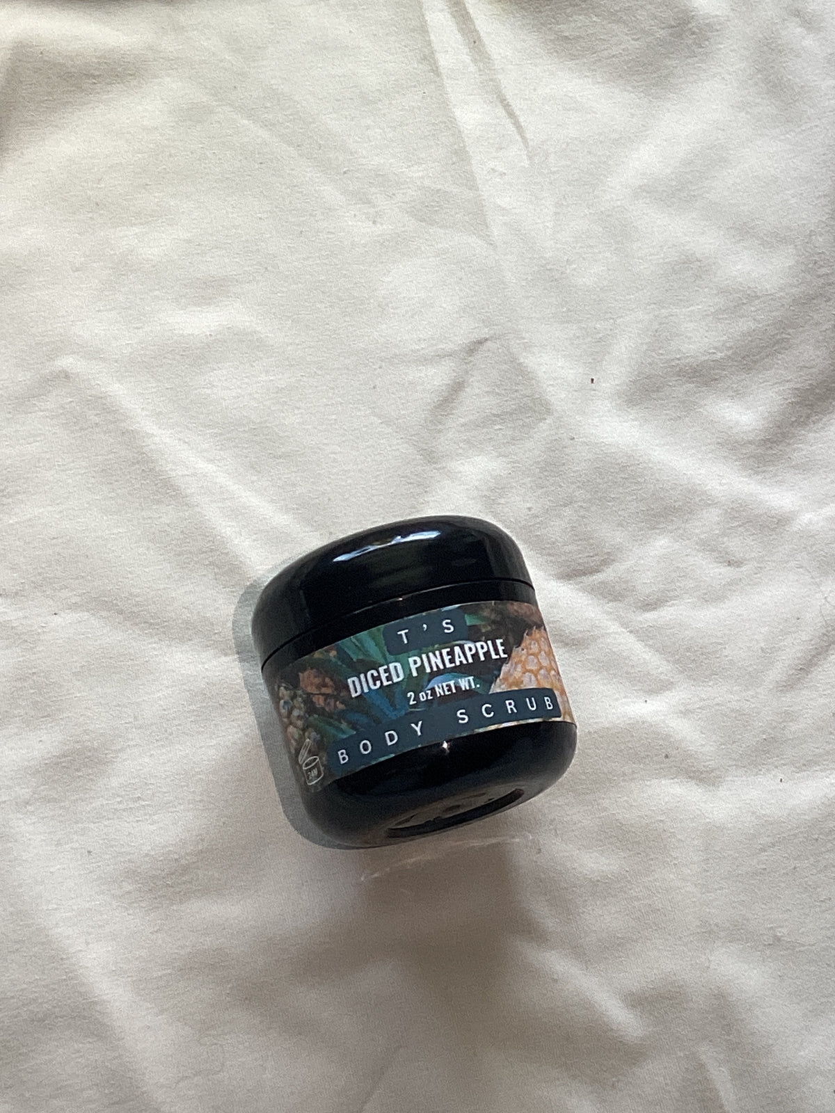A small black plastic jar of body scrub with a decorative label reading 'PINEAPPLE' and 'BODY SCRUB', placed on a wrinkled white fabric background.
