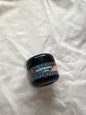 A small black plastic jar of body scrub with a decorative label reading 'PINEAPPLE' and 'BODY SCRUB', placed on a wrinkled white fabric background.