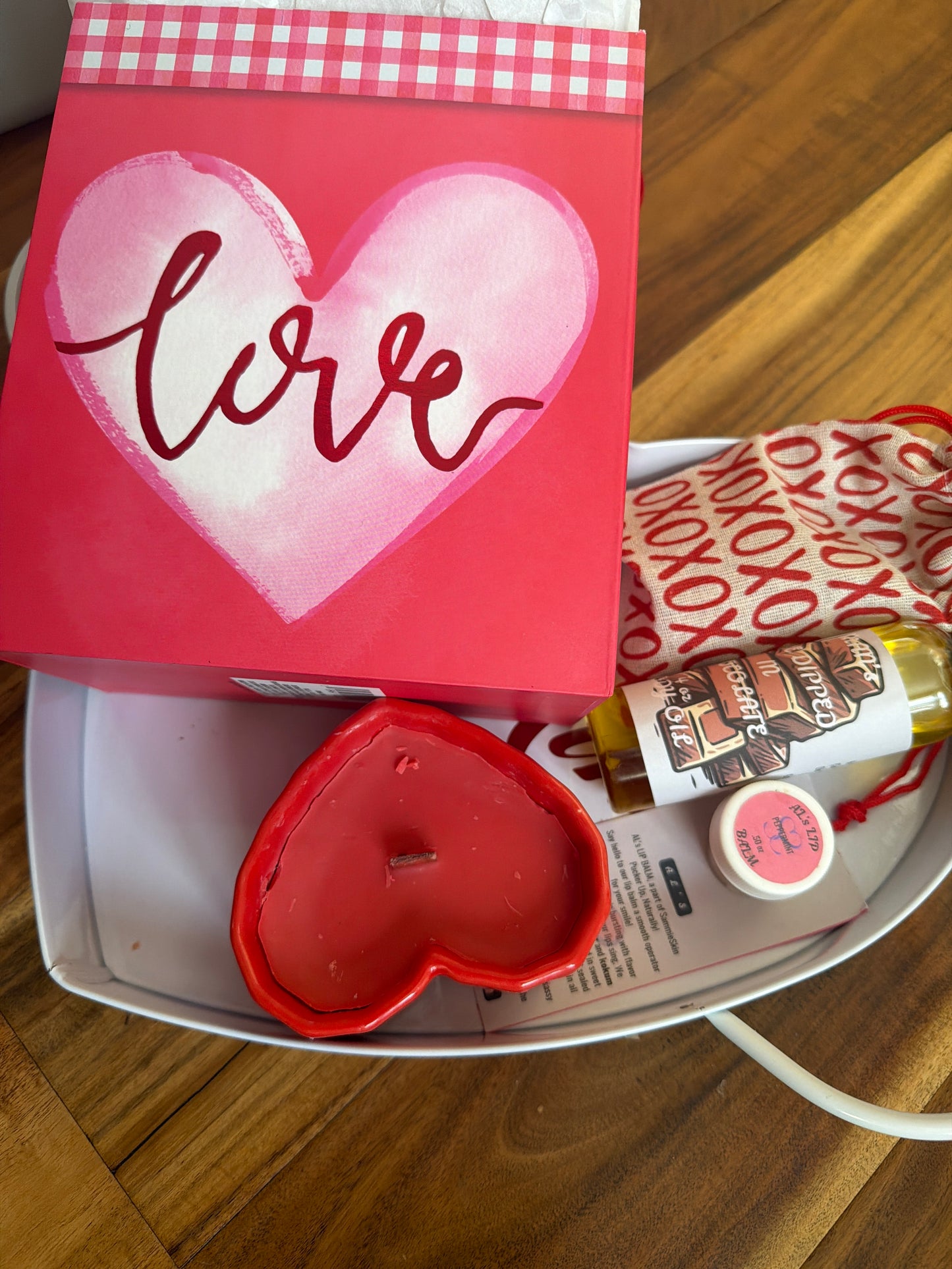 A white tray on a wooden floor with a red candle, body oil squeeze bottle, lip balm on info card and small canvas bag with red Valentine’s Day box.