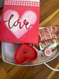 A white tray on a wooden floor with a red candle, body oil squeeze bottle, lip balm on info card and small canvas bag with red Valentine’s Day box.